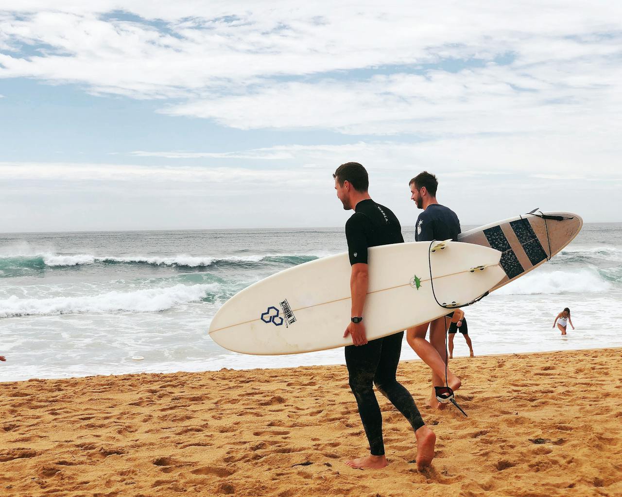 about-me-01 Two men with surfboards walking along Avoca Beach, NSW, capturing the essence of a summer day on the Australian coast.