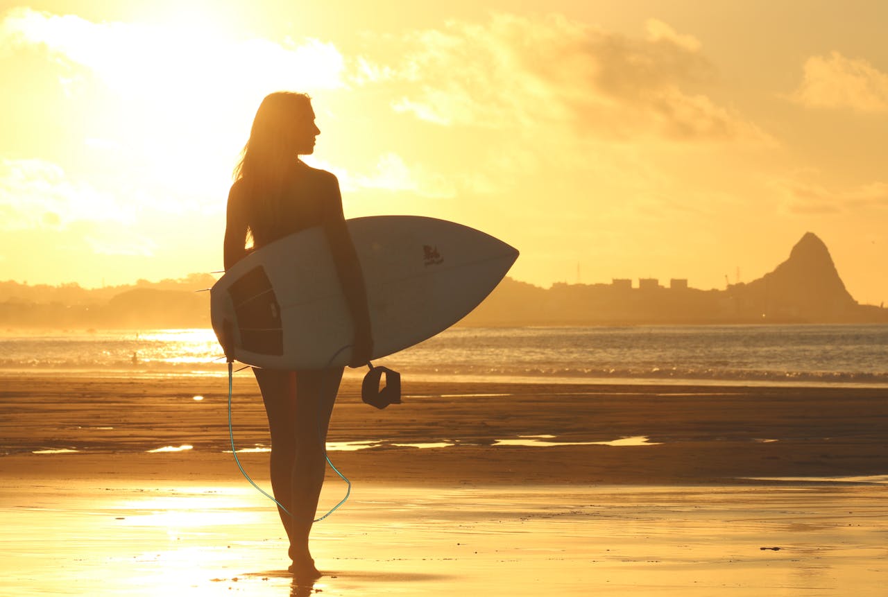 services-01 A silhouette of a woman holding a surfboard, standing on a beach at sunset with a golden sky.